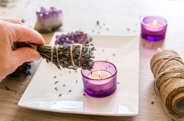 Obraz na płótnie Canvas Person holding homemade herbal lavender (lavendula) smudge stick with smoke coming out, candles and amethyst crystal clusters for decoration. Spiritual home cleansing concept.