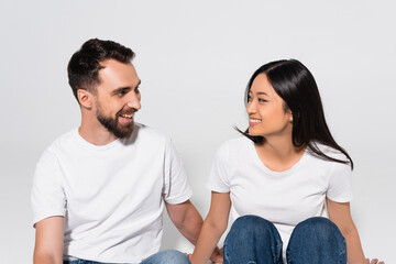 young interracial couple in white t-shirts looking at each other while sitting on white