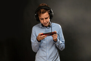 A teenage boy in headphones, intently playing a game on his smartphone. Copy space. Black background. Concept of entertainment and modern technologies