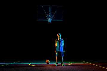 Basketball. A teenager in blue sportswear poses on the Playground, next to a basketball. Dark background, multi-colored neon lighting. Copy space. Concept of sports games