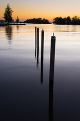 Row of poles in smooth water on the shore of a lake at dusk showing the reflection of sunset in the water a concept of perspective or the fall of the evening