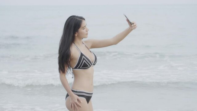 Beautiful young asian woman in bikini sexy taking photo with smartphone on the beach for leisure in summer, girl to travel selfie in vacation with phone for relax on sea in tropical holiday.