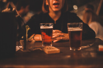 Giovane ragazza seduta al tavolo di un locale serale all'aperto con davanti due bicchieri di birra fresca - obrazy, fototapety, plakaty
