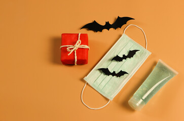Top view  of red gift box with black paper bats , medical face mask and alcohol sanitizer gel  on orange background . Halloween present concept , Covid19 protection and new normal concept.