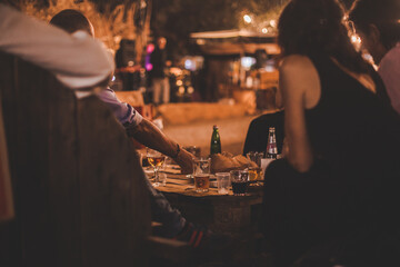 Persone di spalle sedute in un locale serale all'aperto degustano cibo e bevande su tavolo di legno