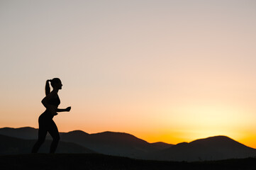 Silhouette against sky at sunrise while running. Runner fitness girl in sport tight clothes. Athlete woman on the road in the mountains. Bright sunset and blurry background. Workout wellness concept.