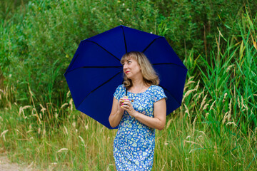 Young blond woman in a blue dress holding a blue umbrella.  The girl is standing in th
