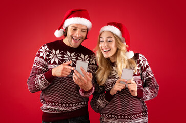 Happy millennial couple in Santa hats shopping for Xmas gifts online on red background