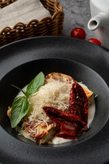 A big piece of italian lasagna in a restaurant, black plate, grey background