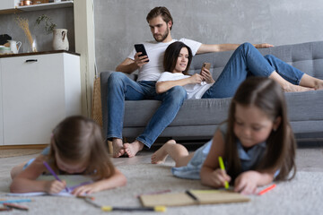 Children sisters playing drawing together on floor while young parents relaxing at home on sofa, little girls having fun, friendship between siblings, family leisure time in living room