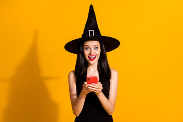 Photo of pretty lovely young magician lady hold telephone excited read holiday post sharing friend liking wear black wizard headwear dress isolated bright yellow color background