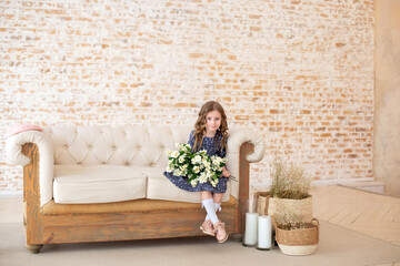 Smiling little curly-haired girl with a bouquet of jasmine flowers sits on the sofa in a cozy...