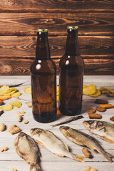 Brown glass bottles of beer and dried fish with chips, nuts, crackers on paper on a white wooden background.