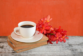 The coffee is placed on an old book with red orchids on the side. The back wall is cement painted orange.