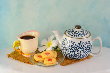 Breakfast is served with tea, in glass and in the pot. There are cookies in the plate and the flowers are placed on a white table. The back wall is a patterned blue cement wall.