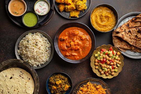 Variety of Indian food, different dishes and snacks on dark rustic background. Pilaf, butter chicken curry, rice, palak paneer, chicken tikka, dal soup, naan bread, assortment of chutney. Top view.