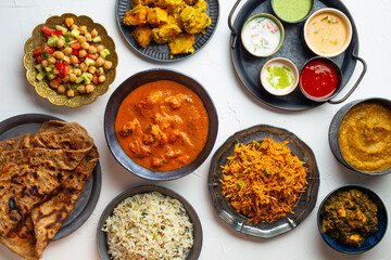 Variety of Indian food, different dishes and snacks on white rustic background. Pilaf, butter chicken curry, rice, palak paneer, chicken tikka, dal soup, naan bread, assortment of chutney. Top view.