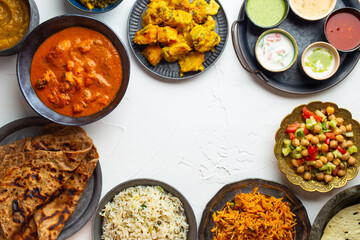 Variety of Indian food, different dishes and snacks on white rustic background. Pilaf, butter chicken curry, rice, palak paneer, chicken tikka, dal soup, naan bread, assortment of chutney. Top view.