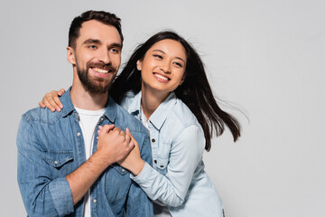 stylish interracial couple in denim shirts holding hands and looking away isolated on grey