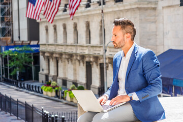 Young European Businessman with beard traveling, working in New York City, wearing blue blazer, sitting on street outside office building, working on laptop computer, looking up, thinking..