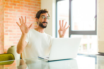 young bearded man with a laptop smiling and looking friendly, showing number nine or ninth with...