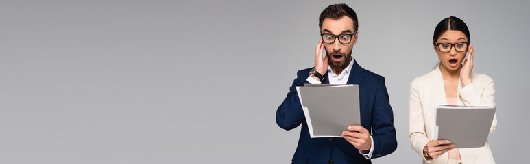 panoramic crop of shocked interracial businesspeople holding hands near faces while looking at...