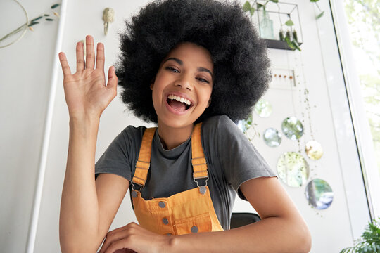 Pretty Young African American Mixed Race Hipster Vlogger Woman With Afro Hair Waving Hand Looking At Webcam Talking To Camera Sits At Cafe Table Video Calling, Recording Blog, Headshot Portrait.