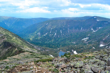 rocks on mountain ridge, view of mountain lake in form of heart among snow ice glaciers, green mountain ranges overgrown with trees, Chersky peak