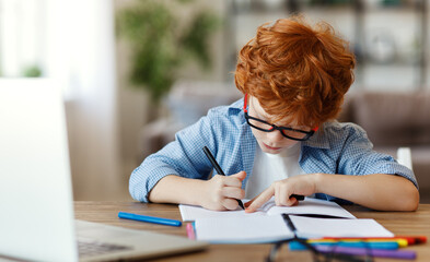 Naklejka na ściany i meble Little schoolboy in glasses doing homework at home.