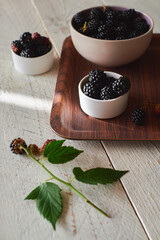 A cup with fresh black and red blackberries on a brown tray, green blackberry leaves is on the table. Light wood background
