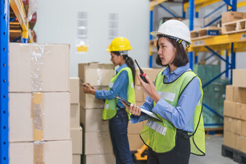 Young Asian woman worker in safety vest with yellow helmet using tablet checking products in stock at warehouse factory