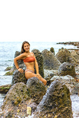 A young girl in a red swimsuit sits on large rocks against the backdrop of the sea.