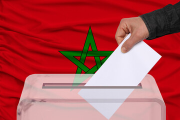 male voter drops a ballot in a transparent ballot box against the Morocco national flag, concept of...