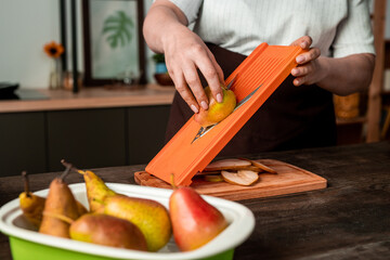 Close-up of unrecognizable woman standing at kitchen counter and slicing pear on mandoline