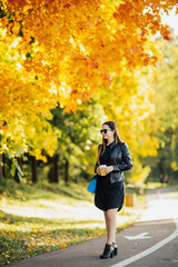 Girl smiling, holding takeaway coffee cup, standing in park in autumn, wearing modern clothes.