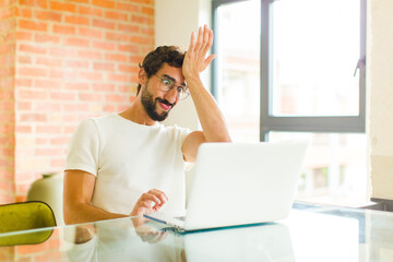 young bearded man with a laptop raising palm to forehead thinking oops, after making a stupid...