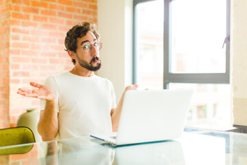 young bearded man with a laptop feeling puzzled and confused, unsure about the correct answer or decision, trying to make a choice