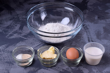 Set of ingredients to make yeast dough. Flour, eggs, butter, dry yeast, sugar and milk in bowls and cups on the grey table