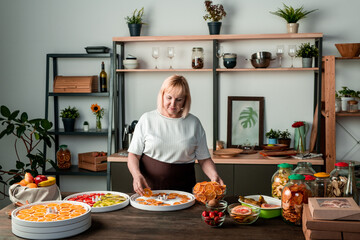 Blond mature woman standing at counter in cozy kitchen and spreading fruit slices on plastic tray while making healthy snacks
