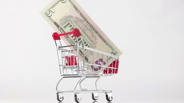 metallic shopping cart of red color with 5 dollar bill rotating against white and gray background, side view. wholesale and black friday sales concept
