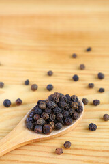 Heap of black peppercorns on wooden spoon with some scattered on wooden table