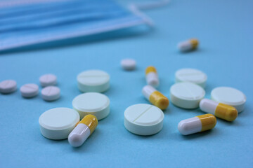 White pills, yellow capsules, and coronavirus medical disposable face mask on blue background with copy space.