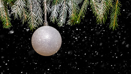 Silver Christmas ball on a snow-covered Christmas tree on a black background during a snowfall