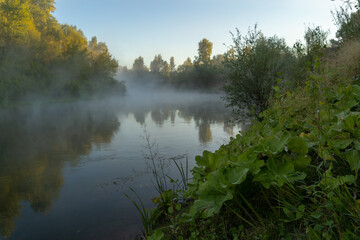 Fototapeta na wymiar Autumn time. Dawn over the river in a misty, brooding haze. Beautiful view of the forest and river, covered with fog in the early morning. The sun's rays of light.
