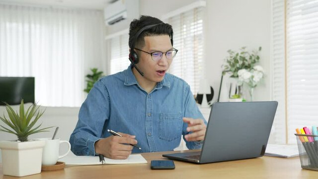 Smiling asian man wear wireless headset making conference videreo call on laptop. Male professional call center agent, hr manager having distance webcam chat job interview on computer at home office.