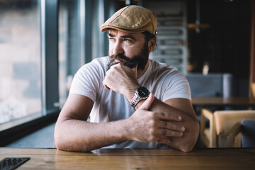 Serious handsome bearded man in casual wear holding hands pondering on ideas and plans for business looking at window, contemplative caucasian mature 40 years male sitting in cafe interior dreaming