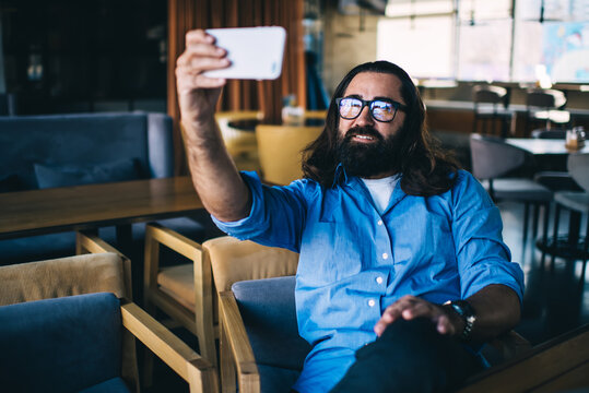 Handsome mature male in casual wear enjoying free time in cafe holding mobile phone for taking selfie, smiling caucasian 40 years man using smartphone for share media content in blog online
