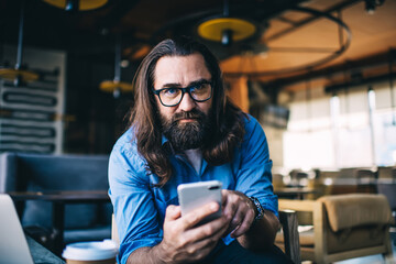 Portrait of handsome bearded mature man in eyewear for vision correction holding mobile phone dialling number, serious confident 40 years male looking at camera using smartphone connect to 4G internet