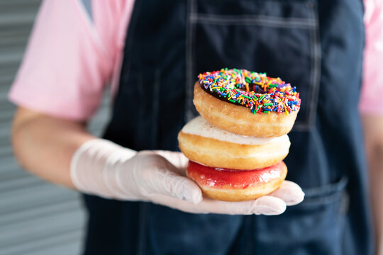 Stacking chocolate frosted donut with sprinkles, sugar-glazed frosted and Bavarian donut on woman hand that wearing latex gloves. Playful and joyful tasty sugary comfort food for customers.