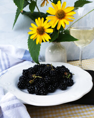 A plate of blackberries with a glass of white wine and yellow flowers in vase on the background